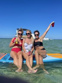 Three friends enjoy a fun day paddleboarding in clear waters, celebrating with drinks and smiles — perfect for a summer getaway.