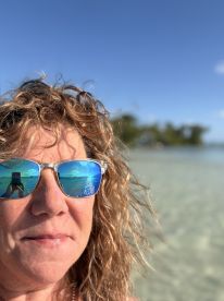 A woman captures a selfie with a stunning beach reflection in her sunglasses, showcasing a perfect day by the water.
