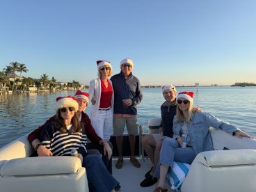 A cheerful group of friends celebrates the holiday season aboard a boat, wearing festive Santa hats against a picturesque waterfront backdrop.