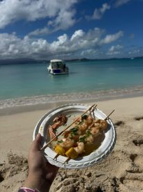 A delicious plate of shrimp skewers enjoyed on the beach, with a boat anchored in the background — perfect for a relaxing day by the water.