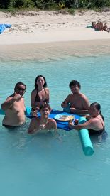 A joyful group enjoys a meal on a floating mat in crystal-clear waters, perfect for a fun day at the beach.