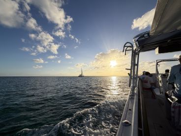 A scenic sunset view from a boat, showcasing a distant sailboat and guests enjoying the ocean experience — perfect for sailing adventures.