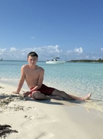 A young teen enjoys a sunny day at the beach, sitting on the sand with a boat visible in the background — a perfect scene for summer adventures.