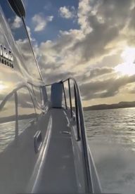 A sleek view of a boat's side as it glides through the water, showcasing the vessel's design against a backdrop of dramatic clouds and a shimmering sea.