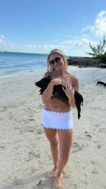 A woman smiles while holding a pig on the beach, showcasing a delightful human-animal interaction in a tropical setting.