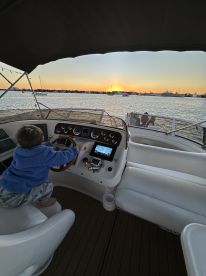 A young child playfully engages with the helm of a boat as the sun sets over the water, capturing a moment of family fun aboard.
