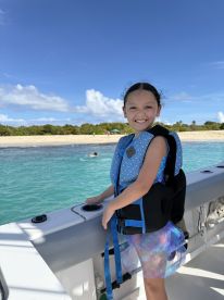 A joyful child in a life jacket smiles on the boat, with a beautiful beach and clear water in the background — perfect for family adventures on the water.