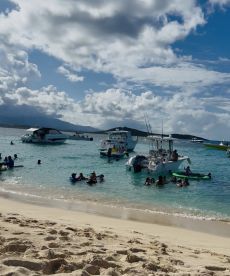 A lively beach scene with groups of people swimming and enjoying the water, surrounded by various boats — perfect for a fun day out on the water.