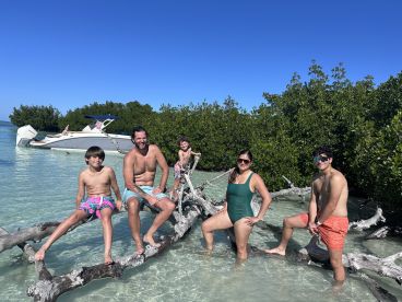 A joyful family enjoys a fun day at the beach, posing on a log in shallow water with their boat anchored nearby — a perfect scene for memorable summer adventures.
