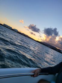 A stunning sunset view from a boat, with a hand gripping the railing, capturing the serene beauty of the water and distant shoreline.