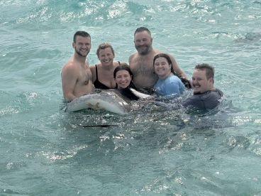 A group of six people joyfully interacts with a dolphin in shallow waters, creating a memorable wildlife experience in a tropical paradise.