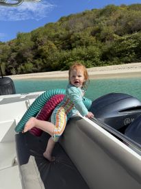 A young child enjoys a sunny day on a boat, playfully posing with colorful pool floats against a beautiful beach backdrop — perfect for family adventures on the water.