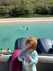 A young child enjoys a refreshing drink while watching friends swim in the clear turquoise waters near a sandy beach, capturing the essence of family fun on a boating adventure.