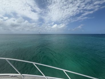 A serene view from the bow of a boat, showcasing the tranquil turquoise waters and a distant sailboat under a partly cloudy sky — perfect for a peaceful day on the water.