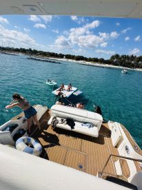 A lively group enjoys a sunny day on a pontoon boat, with some swimming and relaxing on a floating mat near the beach — perfect for a fun day out on the water.