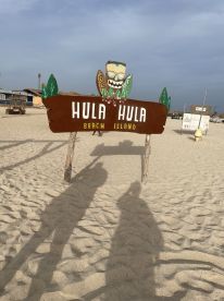 A sign for Hula Hula Beach Island is displayed on a sandy beach, but the image lacks clarity and detail.