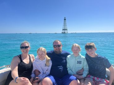 A cheerful family poses on a boat with a lighthouse in the background, enjoying a sunny day on the ocean — perfect for family outings and adventures.