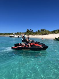 A couple enjoys a thrilling ride on a jet ski in crystal-clear waters, surrounded by a beautiful beach — perfect for adventure seekers and water sports enthusiasts.