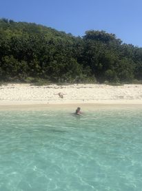 A child enjoys swimming in crystal-clear waters near a sandy beach, while another person relaxes on the shore — a perfect family day at the beach.