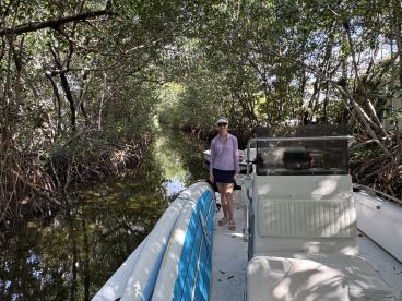 A woman stands on a boat navigating through a serene mangrove channel, showcasing the beauty of nature and exploration on the water.