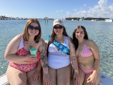 Three friends celebrate a bachelorette party on a sunny day by the water, enjoying drinks and laughter in a picturesque setting.