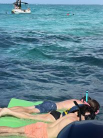 A group enjoys snorkeling and relaxing on a floating mat in crystal-clear waters, with a pontoon boat anchored nearby — a perfect day of fun in the sun.