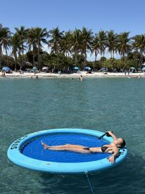 A woman relaxes on a floating mat in clear waters, enjoying a sunny day at the beach surrounded by palm trees — a perfect scene for summer fun and relaxation.