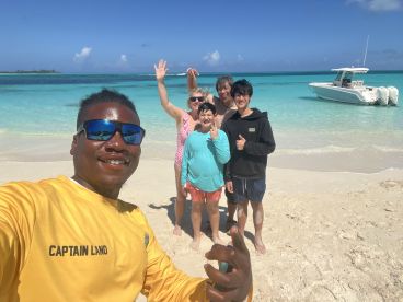 A joyful group poses for a selfie on the beach, with a boat anchored in the background — capturing the essence of a fun day on the water.