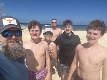 A cheerful family poses for a selfie on the beach, capturing a fun day in the sun with clear blue skies and ocean waves in the background.
