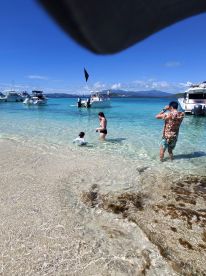A family enjoys a sunny day at the beach, with boats anchored nearby and children playing in the shallow water — a perfect scene for summer fun and relaxation.