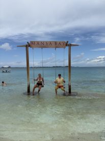 A couple enjoys swinging in shallow waters at Mansa Bay, creating a perfect beach moment filled with fun and relaxation.