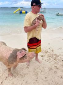 A man holds a piglet while a larger pig stands nearby on the beach, showcasing a unique and playful interaction with wildlife in a tropical setting.