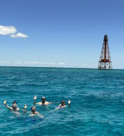 A group of friends enjoys snorkeling in vibrant turquoise waters, waving at the camera with a historic lighthouse in the background — a perfect snapshot of adventure and fun on the water.