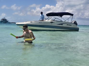 A man enjoys a playful moment in the shallow waters, holding colorful pool noodles while a boat is anchored nearby — perfect for a fun day on the water.