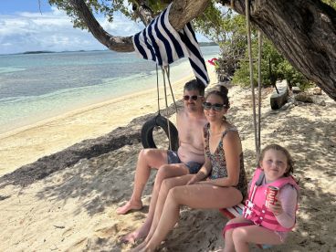 A family enjoys a relaxing day at the beach, sitting on a swing under a tree, with clear waters in the background — perfect for family outings and beach fun.