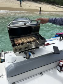 A grill is set up on a boat, cooking skewers while guests enjoy the beach in the background — perfect for onboard barbecues and culinary experiences at sea.