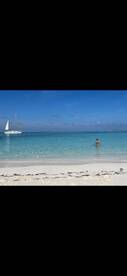 A person enjoys a refreshing swim in the crystal-clear waters of a tropical beach, with a sailboat gracefully anchored nearby — a perfect scene for island relaxation.