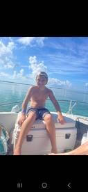 A young boy enjoys a sunny day on a boat, smiling against a backdrop of clear blue waters and skies — a perfect moment of fun on the ocean.