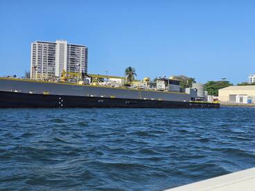 A clear view of a large vessel docked near a waterfront, showcasing its structure against a backdrop of buildings and palm trees.