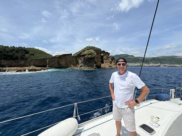 A smiling individual poses on the deck of a sailboat with stunning coastal scenery in the background, capturing the essence of a memorable boating adventure.