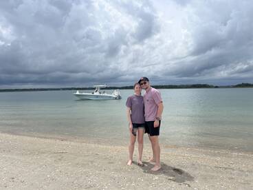 A couple poses on the beach with a boat anchored in the background, capturing a perfect moment of relaxation and adventure by the water.