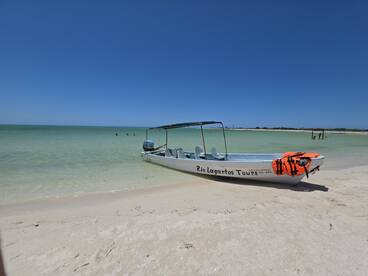 A small tour boat named 'Rio Lagartos Tours' is anchored on a beautiful beach, with guests enjoying the clear waters nearby — perfect for a day of exploration and adventure.