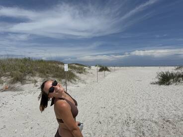 A woman smiles brightly on a sandy beach under a clear blue sky, capturing the essence of a sunny day at the shore.