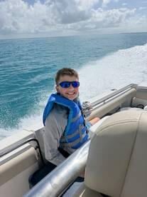 A young boy enjoys a thrilling boat ride, wearing a life jacket and smiling against the backdrop of sparkling blue waters — a perfect moment for family adventures on the sea.