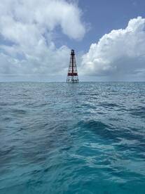 A distant view of a lighthouse standing tall in the ocean, surrounded by turquoise waters and a cloudy sky.