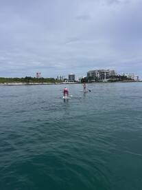 Two individuals enjoy paddleboarding in calm waters, with a scenic coastline and buildings in the background — a perfect day for water sports.