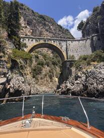 A stunning view of a boat navigating through a narrow passageway beneath a historic bridge, surrounded by rugged cliffs and lush greenery — a picturesque moment on the water.