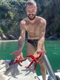 A man joyfully holds two vibrant red starfish, showcasing a unique human-animal interaction in a beautiful marine setting.