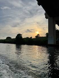 A serene sunset view over calm waters, framed by lush greenery and a bridge, creating a picturesque scene perfect for evening boat rides.