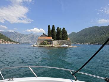 A picturesque view of a small island with lush trees and a historic building, set against a backdrop of stunning mountains and clear blue skies — a perfect destination for boat tours.